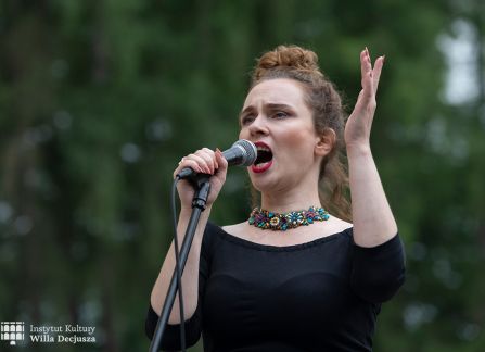 fotografia. dni wyszehradzkie w willi decjusza. koncert zespołu balkan artz. kobieta śpiewa do mikrofonu. w tle nieostre drzewa, zieleń.